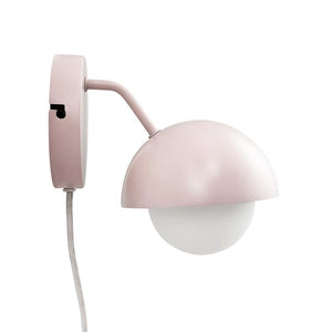 Væglampe i pudderrosa hænger monteret på rund vægbeslag; en halvkugleformet skærm dækker en hvid opalglas-kugle, med buet arm og stofindhyllet ledning mod hvid baggrund.