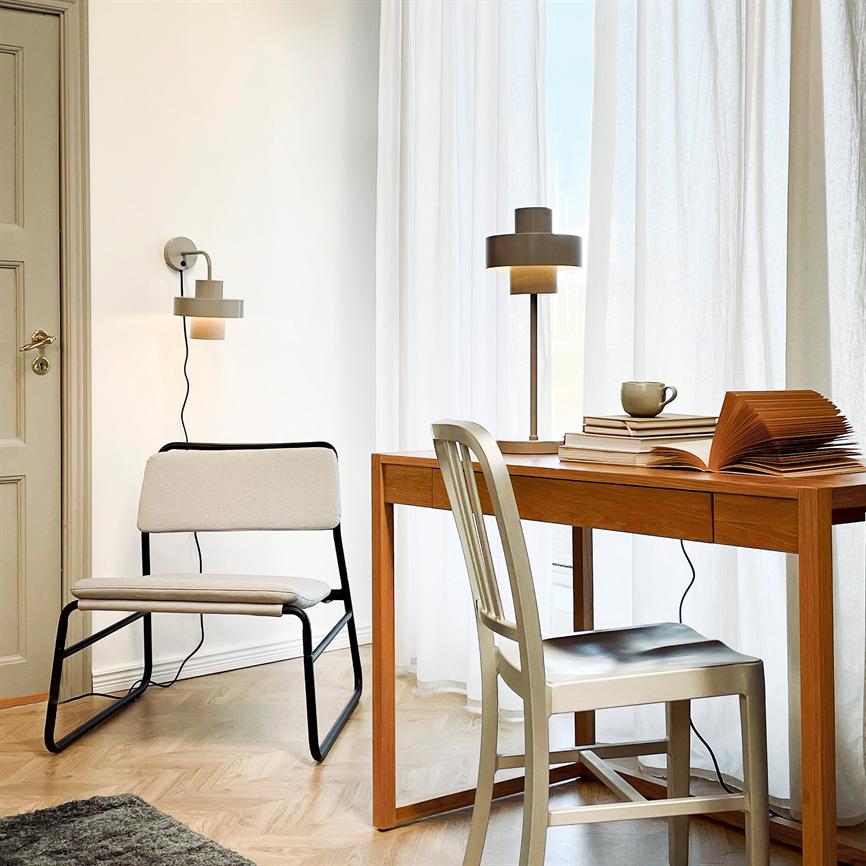 Træskrivebordet står ved et lyst vindue; en åben bog og en kop ligger på bordet, bordlampe lyser diskret; en metalstol foran bordet og en polstret loungestol ved væggen.