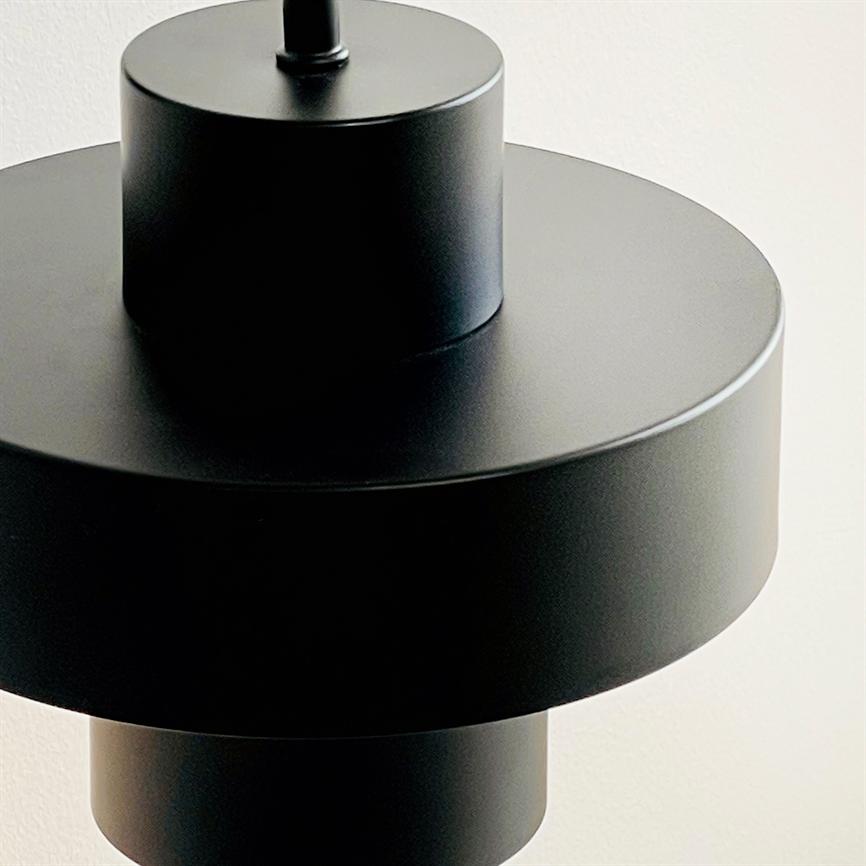 En sort, cylindrisk pendellampe hænger foran en lys væg, bestående af tre stablede matte metaldele med bløde skygger og glatte kanter.