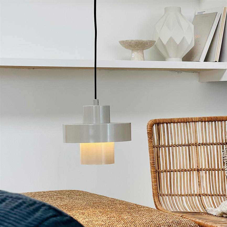 En hvid metalpendel lyser svagt, hænger i sort ledning over en flettet ratanstoleryg og naturfarvet bænk, med en hvid hylde og dekorative vaser bagved.