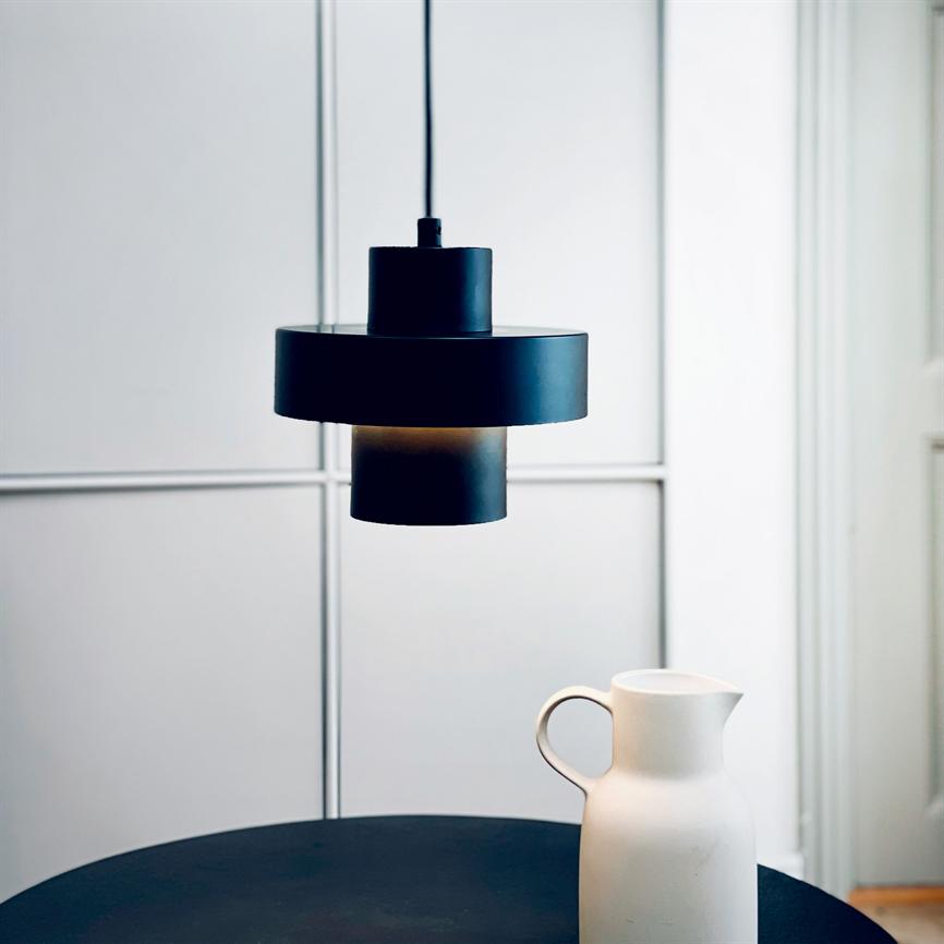 Sort pendel-lampe hænger og kaster blødt lys over et rundt mørkt bord, hvor en hvid keramikkande står; minimalistisk, lyst rum med opdelte vægpaneler i baggrunden.