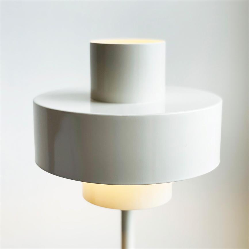 Moderne hvid cylindrisk lampe, to stakede skærme, udsender blødt varmt lys nederst; står på en slank stang foran en lys, neutral væg i et minimalistisk rum.