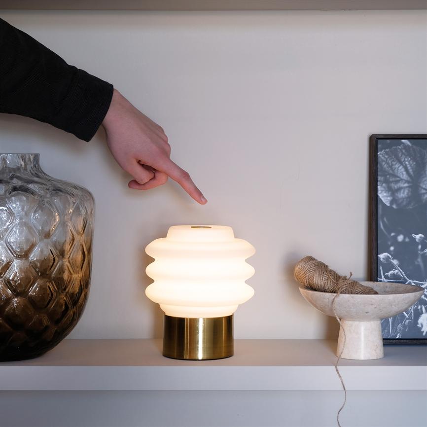 Bordlampe med bølget hvid skærm lyser blødt; en hånd peger mod den; står på en hylde mellem en stor glasvase og en skål med snor, ved siden af en sort-hvid plakat.