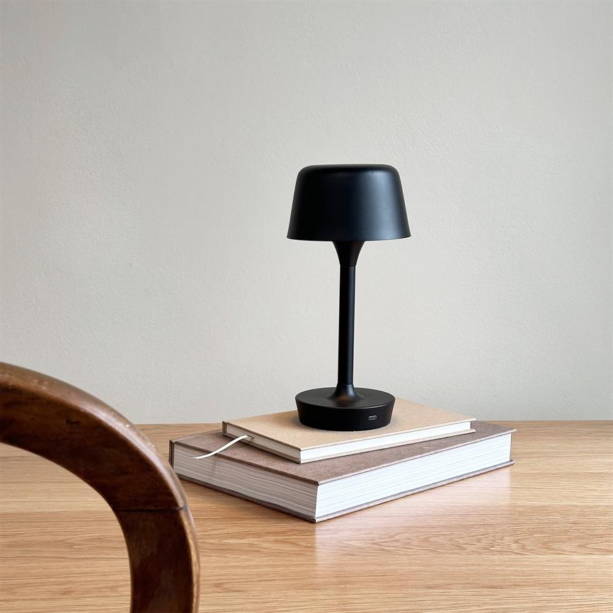 En sort bordlampe står på to stabel- bøger på et lyst træbord foran en ensfarvet væg; en buet, mørk træstol ses delvist i forgrunden til venstre.