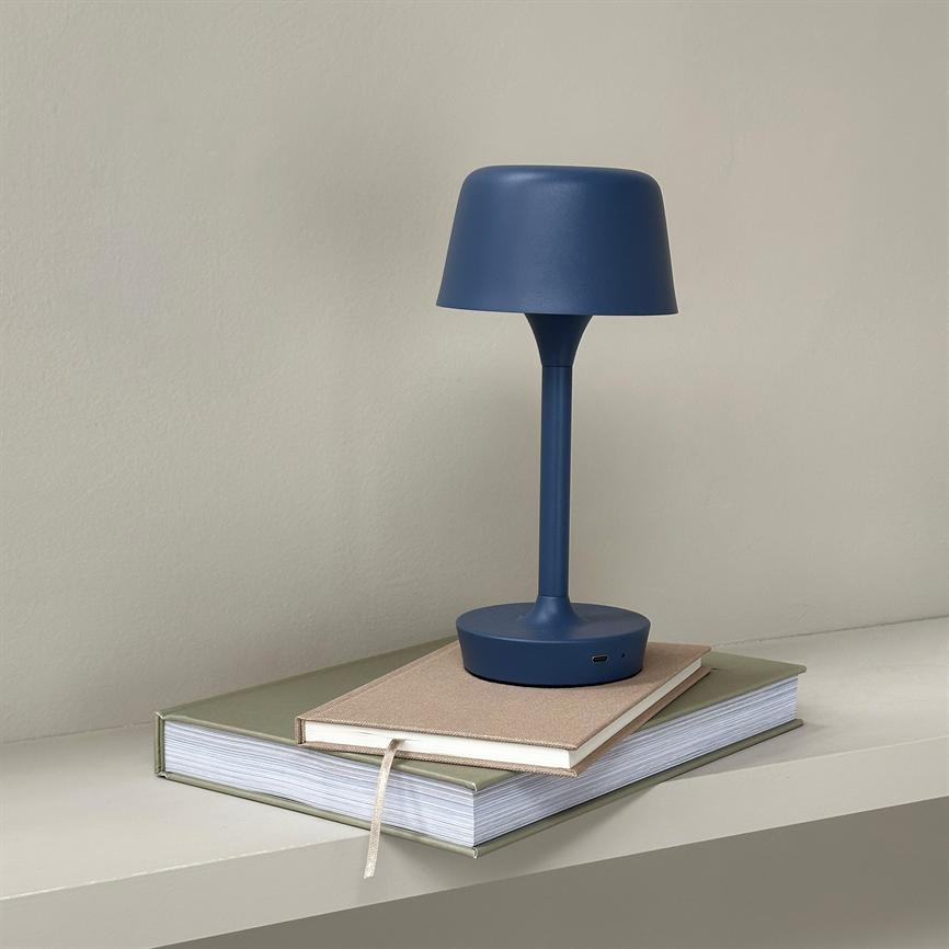 En blå bordlampe hviler på to stablede bøger på en minimalistisk lysegrå hylde, foran en beige væg — enkel, moderne scene med bløde skygger.