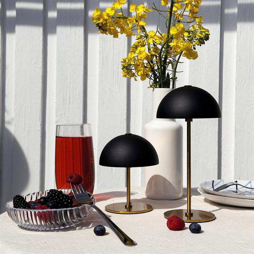 To sorte kuppellamper med messingsokler står på et solbelyst hvidt bord; foran en skål med bær og et glas rød drik, bagved en hvid vase med gule blomster.