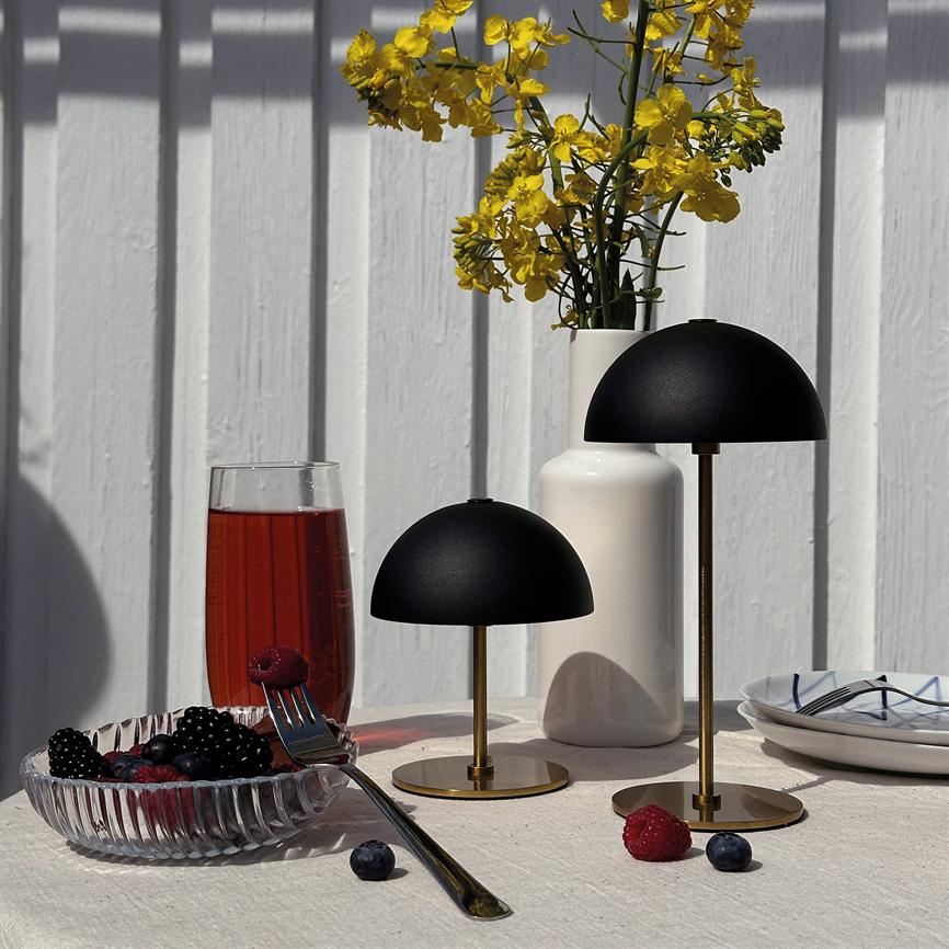 To sorte, svampeformede bordlamper står på et bord ved en hvid trævæg; ved siden af en hvid vase med gule blomster, et glas rød drik og en skål med bær.