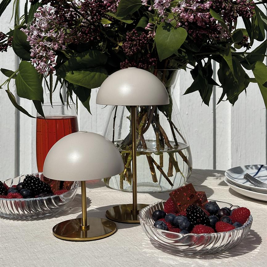 To små hvide kuppellamper står på messingsokler på en hvid dug; omgivet af en glasvase med lilla syrener, skåle med bær og chokolade samt et glas rød drik foran hvid trævæg.