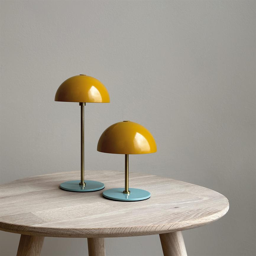 To gule bordlamper står på et rundt, lyst træbord — én høj, én lav — med blågrønne metalfødder foran en mat, lysgrå væg i et enkelt, minimalistisk rum.