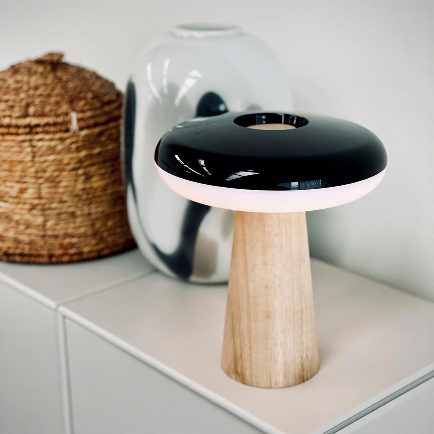 Bordlampe med træfod og rund, sort glossy skærm udsender blødt rosa lys; står på hvid kommode foran en sølvglans vase og flettet kurv i et lyst, minimalistisk rum.