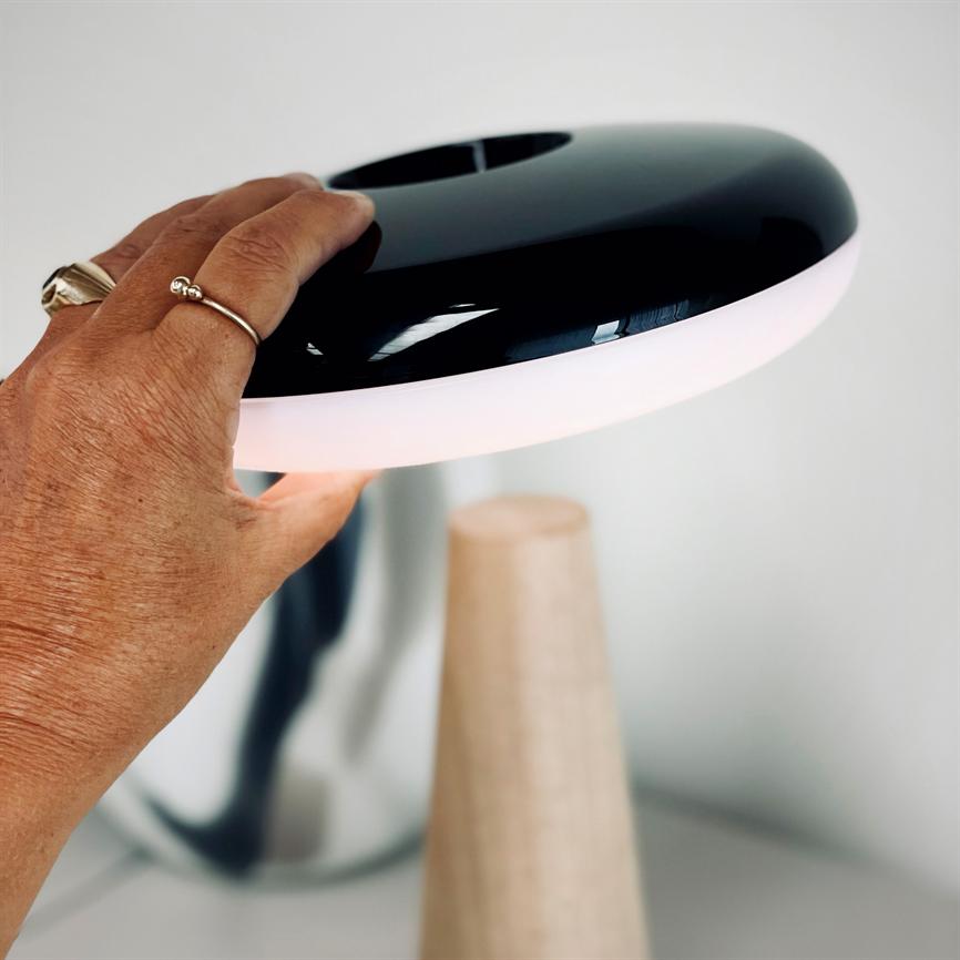 Bordlampe — en hånd løfter den sorte, runde skærm med lysende hvid kant; lampen står på en kegleformet træfod i et lyst, minimalistisk rum.