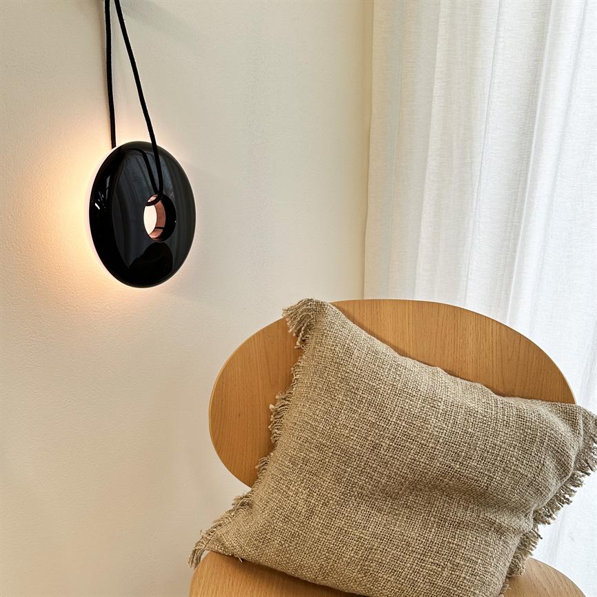 Sort væglampe hænger i sort ledning og udsender varmt baggrundslys; lys træstol med grov beige pude står foran hvide vægge og et hvidt gardin i et roligt hjørne.