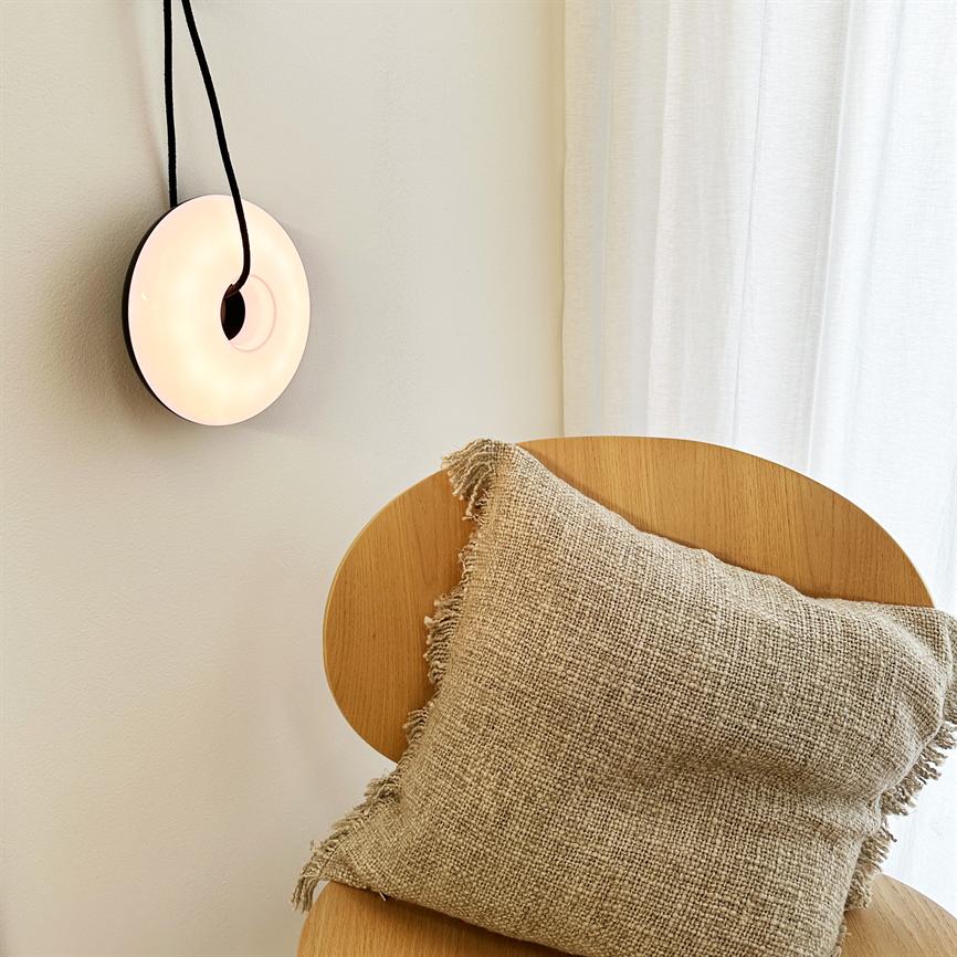 En rund væglampe hænger og udsender blødt lys; en beige, grovvævet pude hviler på en ovalrygget træstol i et lyst rum med cremefarvet væg og hvide gardiner.