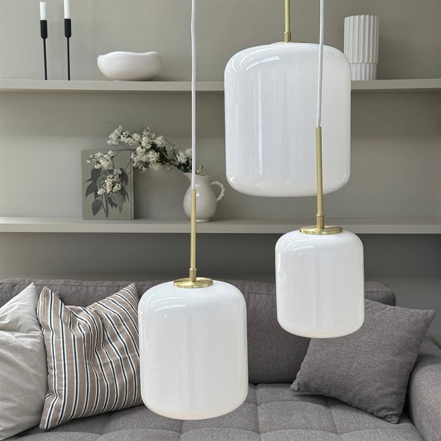 Tre hvide pendellamper hænger over en grå, polstret sofa; foran hylder med en vase med hvide blomster, dekorative skåle og stearinlys i et roligt, neutralt indre.