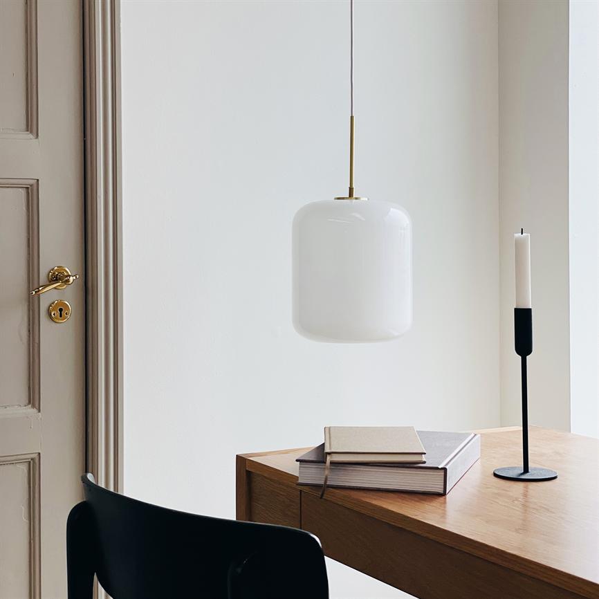 En hvid glaspendel hænger over et træbord; på bordet ligger en stak bøger og en sort lysestage med et hvidt stearinlys, en sort stol står ved en dør med messinghåndtag.