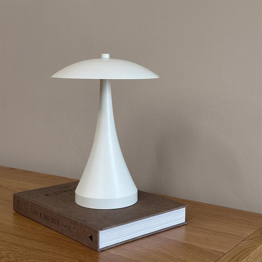 Hvid, svampformet bordlampe står på en tyk, brun bog placeret på et lyst træbord foran en ensfarvet beige væg. 
 Tekst på bogryg: THE KINFOLK TABLE.