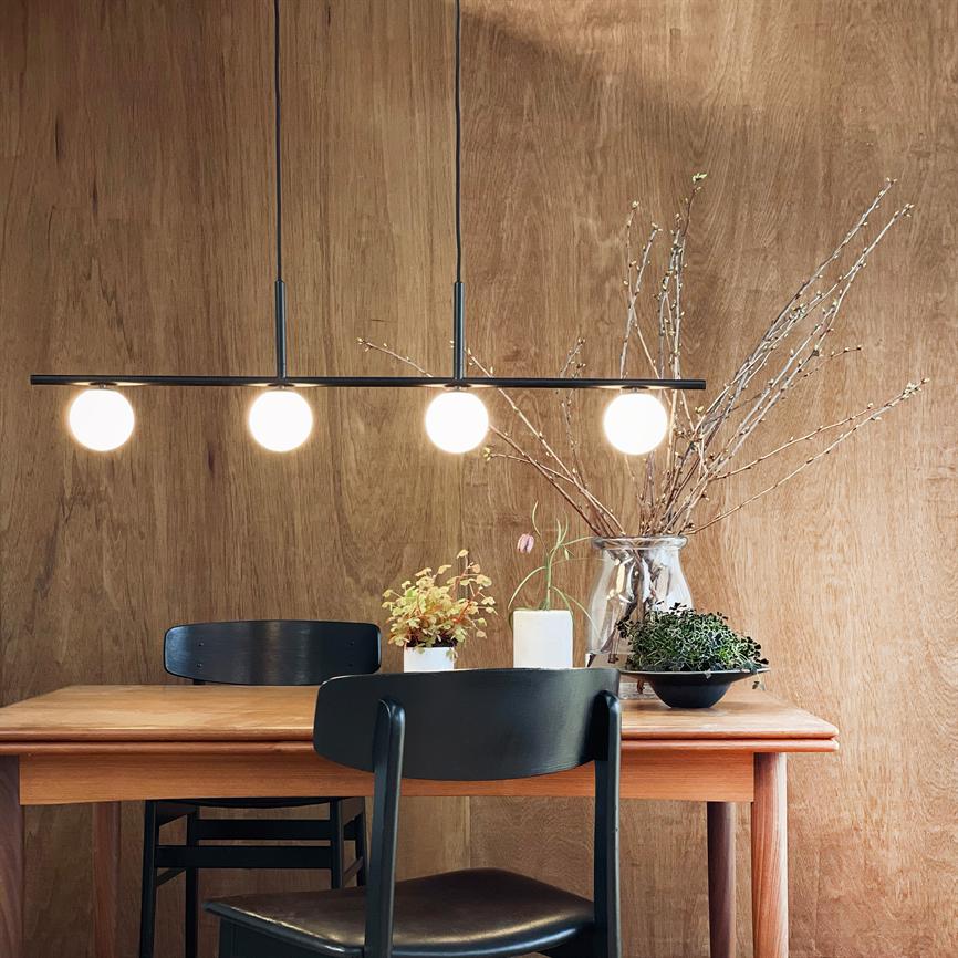 Pendelarmatur med fire runde, tændte pærer lyser over et træspisebord, omgivet af sorte stole, små planter og en glasvase med grene foran en træbeklædt væg.