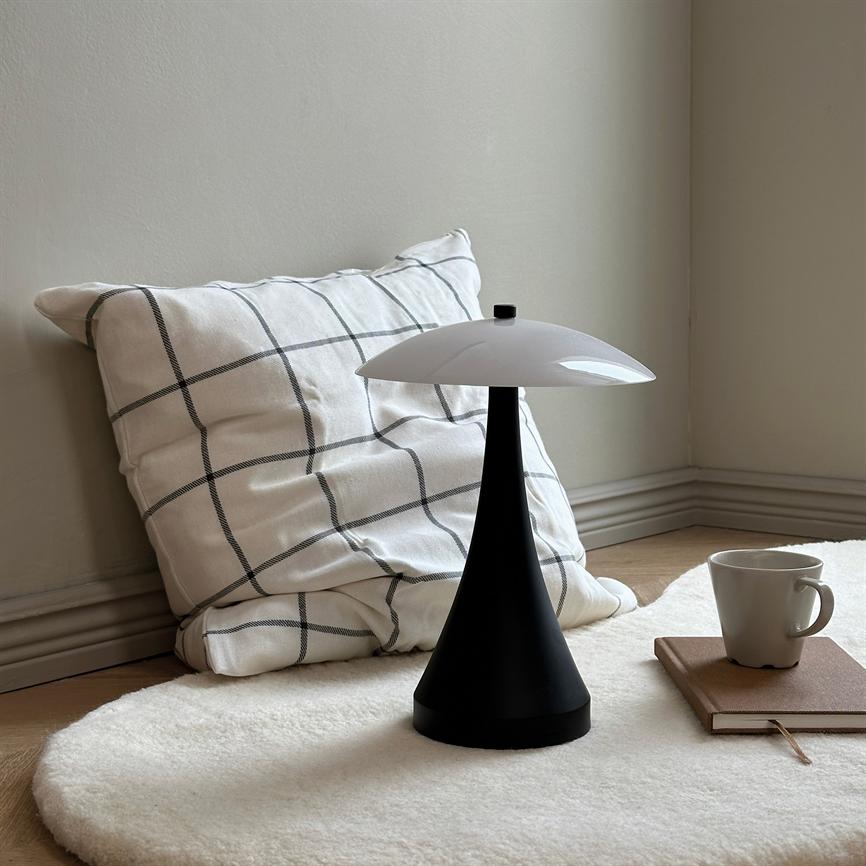 En sort, kegleformet bordlampe med hvid kuppel står midt på et blødt rundt hvidt tæppe; en ternet pude læner sig mod væggen, ved siden ligger notesbog og en kaffekop.