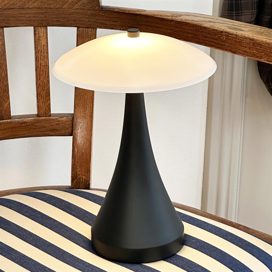 En sort kegleformet bordlampe udsender varmt lys under en hvid kuppel, placeret på en blå-hvidstribet hynde på en træstol i et lyst, indendørs hjørne.