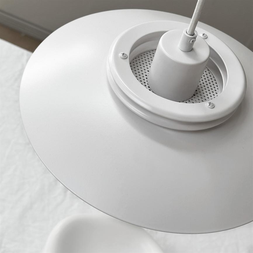 En hvid metal pendellampe hænger fra en stofbeklædt ledning; lampens runde skærm og perforerede fatning ses skråt. Kontekst: lyst, minimalistisk interiør med hvidt tekstil og neutral væg.
