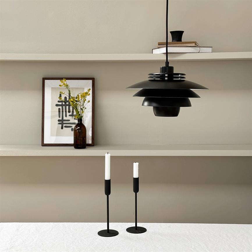 En sort, lagdelt pendellampe hænger over et hvidt dækket bord; to slanke sorte lysestager med hvide stearinlys står på bordet. I baggrunden hylder med en indrammet plakat og en brun flaske med gule blomster.