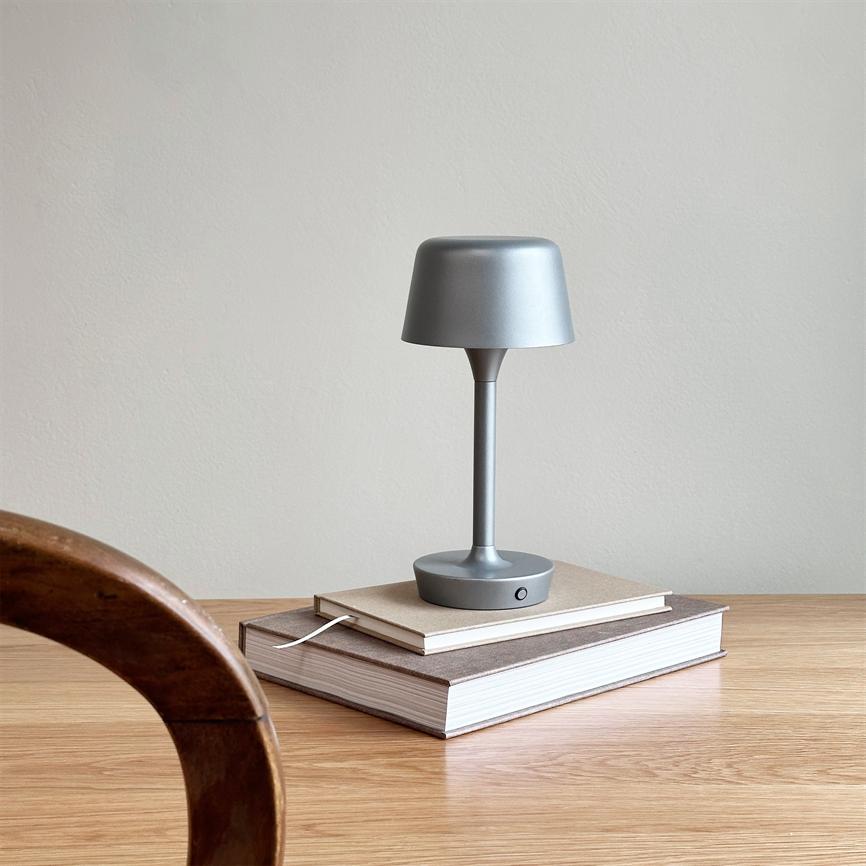 En lille grå bordlampe står på to stablede bøger på et lyst træbord; en buet trækant af en stol i forgrunden og en neutral væg i baggrunden.