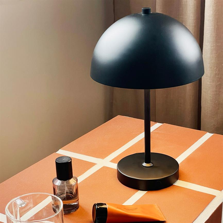 Sort bordlampe står på et orange bord med hvide fliselinjer, omgivet af et glas, en parfumeflakon og en orange tube; beige gardin og væg i baggrunden.