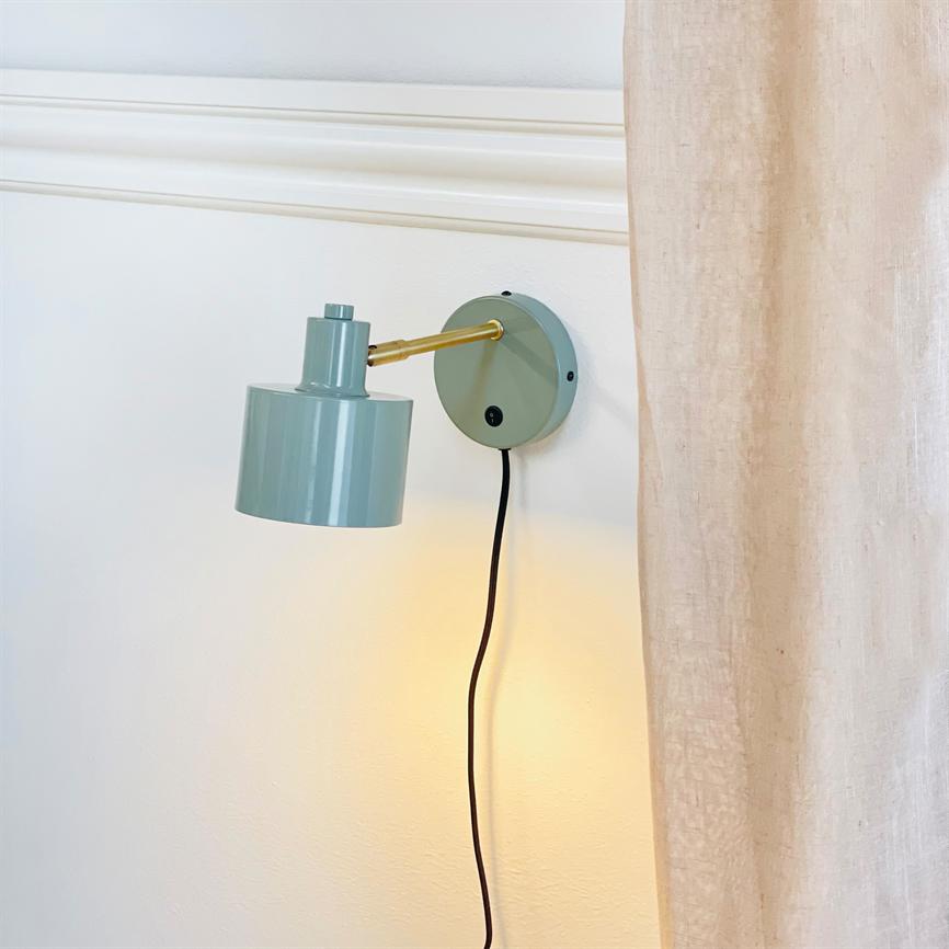 Væglampe i mintgrønt, monteret på en rund vægplade, kaster varmt lys nedad; guldfarvet arm og sort ledning hænger mod en hvid væg ved et beige gardin.