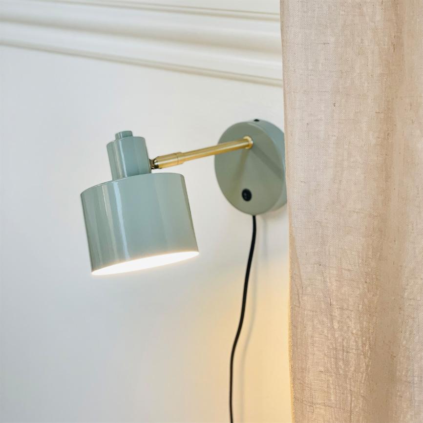 En mintgrøn væglampe monteret på en rund vægplade, arm i messing; lampens skærm lyser nedad. Placeret mod hvid væg med synlig sort ledning og beige gardin til højre.