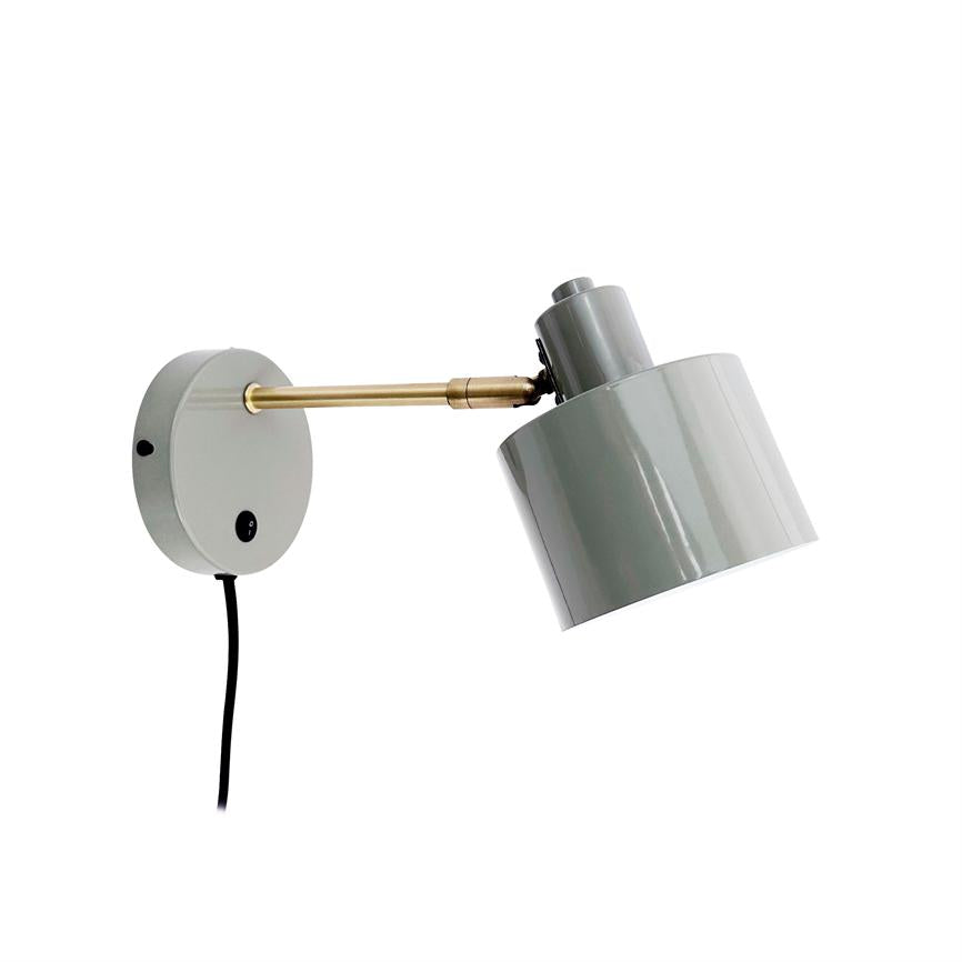 Væglampe monteret på rundt vægbeslag; messingarm strækker sig ud, sort ledning hænger ned, cylinderformet grå skærm er vinklet nedad mod hvid baggrund.