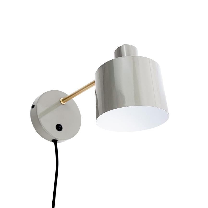 Grå cylinderformet væglampe med guldfarvet arm monteret på rundt vægbeslag, en sort ledning hænger ned; lampeskærmen peger nedad mod en neutral, hvid baggrund.