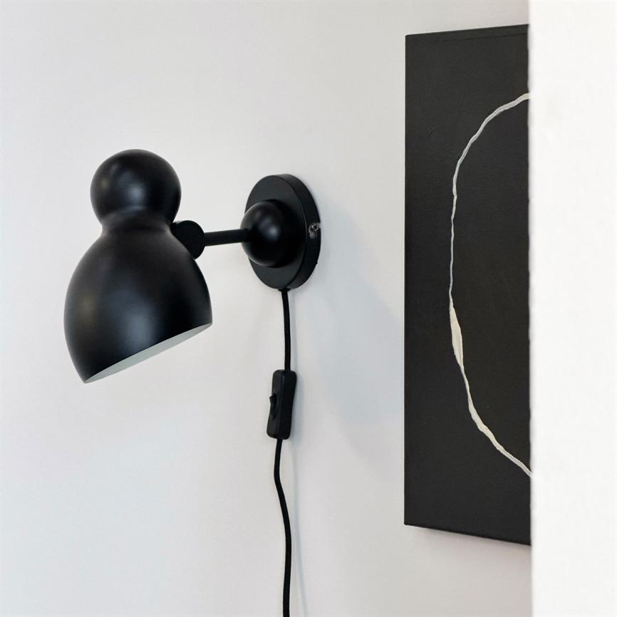 En sort væglampe sidder monteret på en hvid væg, dens arm peger skråt nedad, ledning med afbryder hænger frit; ved siden af et sort maleri med en brudt hvid cirkel.