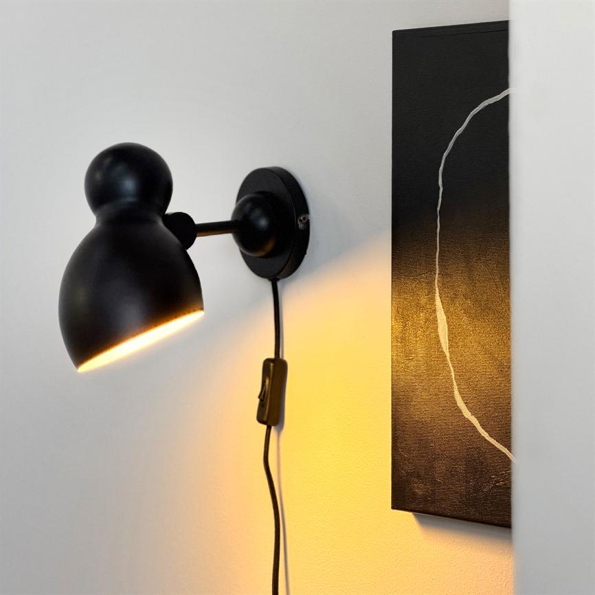 Sort væglampe lyser varmt nedad, kaster gulligt skær på en hvid væg ved siden af et smalt sort maleri med en tynd, hvid buet linje.