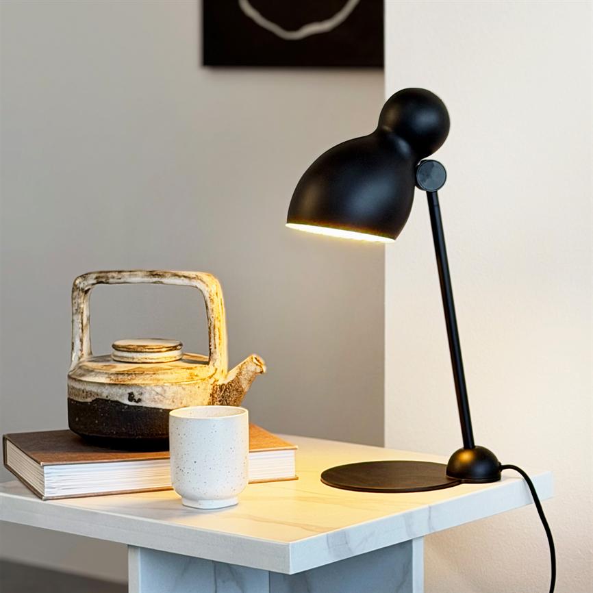 Sort bordlampe lyser over en hvid kop og en rustik tekande placeret på en bog på et lyst natbord i et roligt, minimalistisk rum.