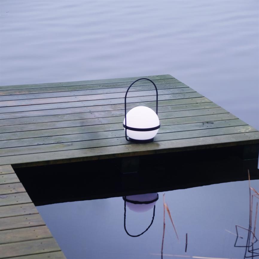 En rund, oplyst lanterne med sort bøjle hviler på en træbrygge og kaster sit spejlbillede i det rolige søvand.