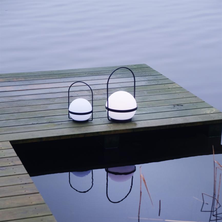 To runde, hvide lanterner i metalbøjler står og udsender blødt lys på en slidt træmole ved en stille sø, deres refleksioner synlige i det rolige vand.