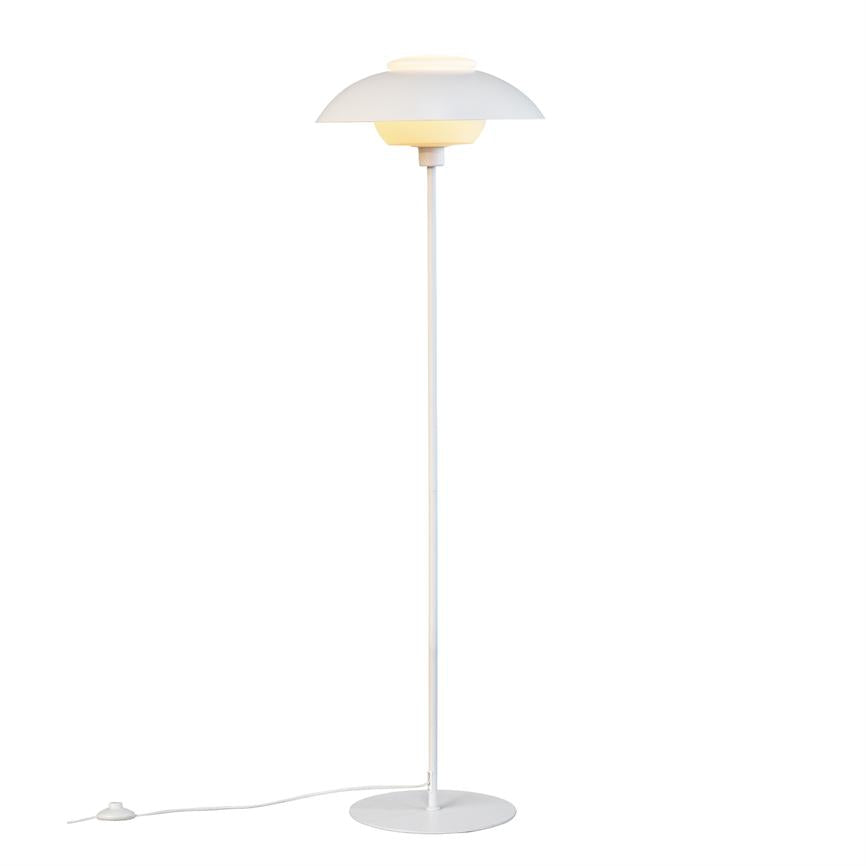 Hvid gulvlampe står tændt; rund skærm udsender varmt lys, slank stang forbinder skærm med rund fod, strømkabel med fodkontakt ligger udstrakt på hvid baggrund.