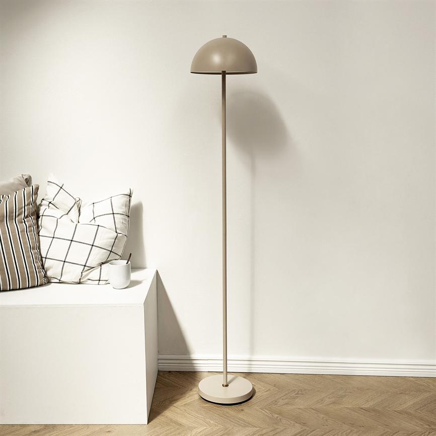 Gulvlampe med halvkugleformet skærm står oprejst ved en lav hvid bænk med ternet og stribet pude samt en hvid kop, i et minimalistisk hjørne med lyst trægulv og hvid væg.