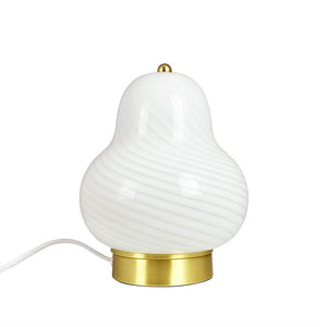 Hvid pæreformet bordlampe med bølgende rillede glasoverflade, med lille guldknap øverst, placeret på rund messingfod; elektrisk ledning løber ud til venstre mod en ensartet hvid baggrund.
