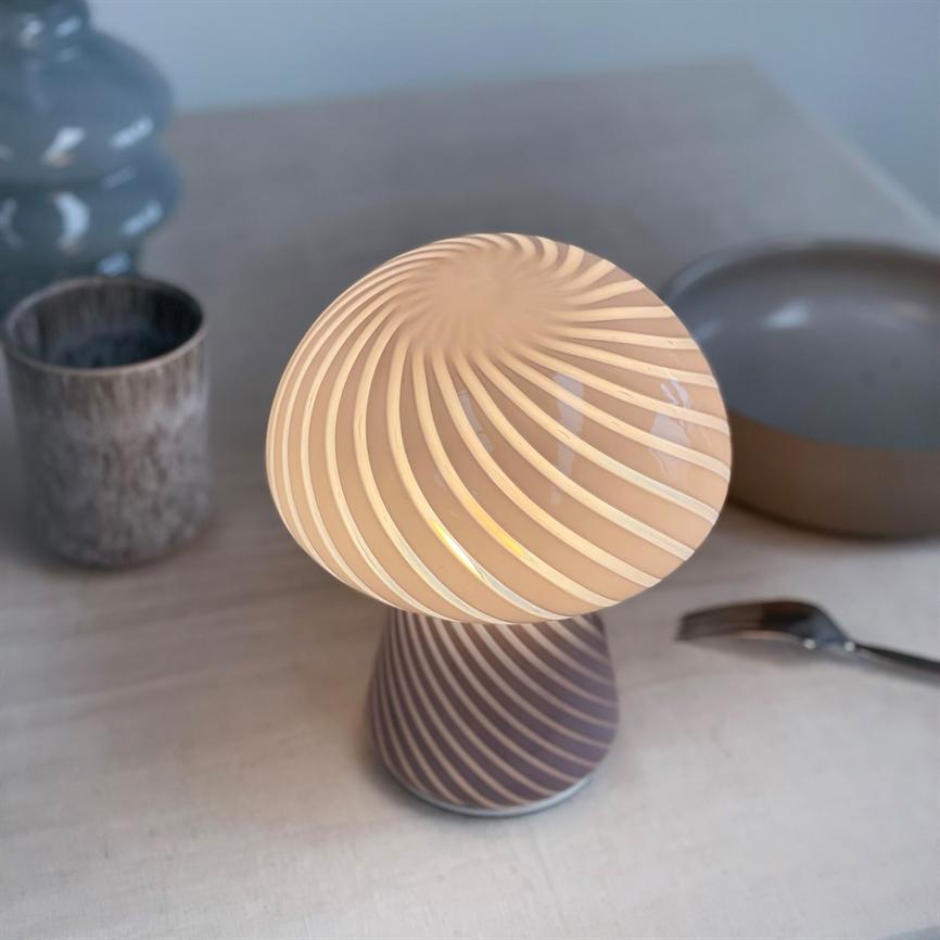 En lille spiralformet bordlampe udsender varmt, diffust lys og står på et lyst bord ved siden af en grå skål, en rustik kop og en gaffel.