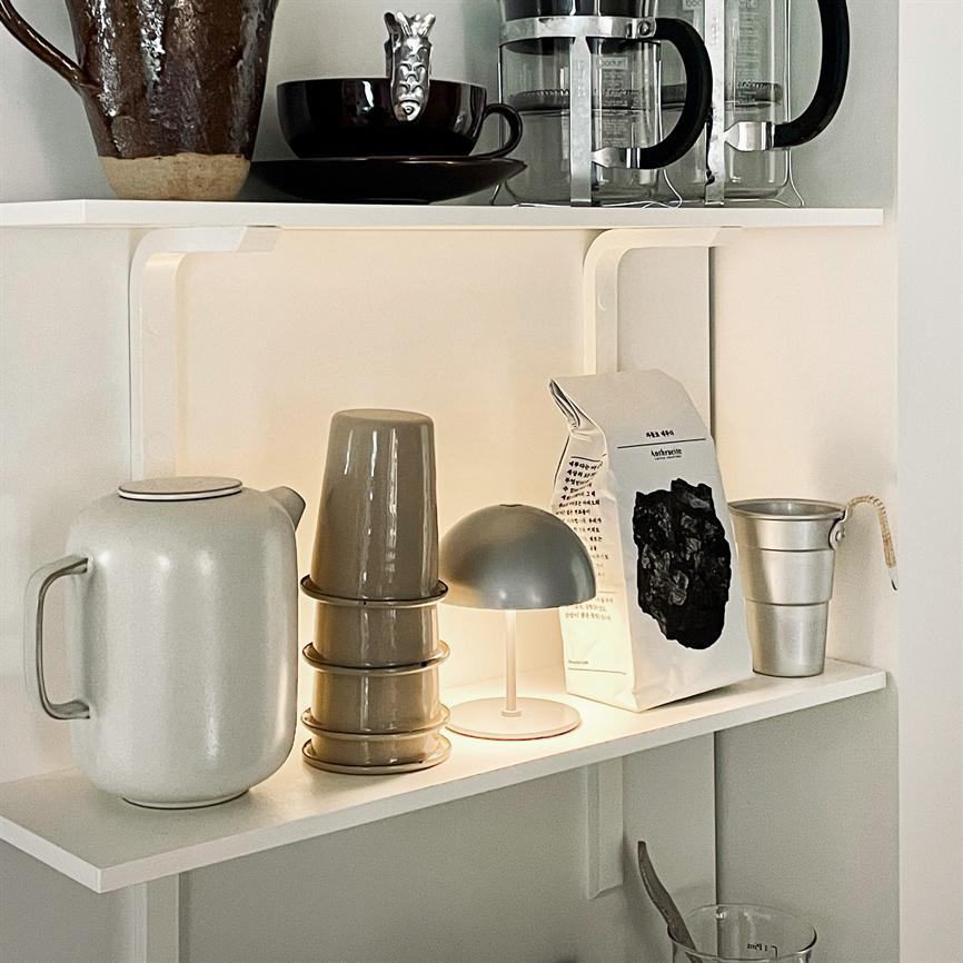 En lille grå bordlampe lyser svagt på en hvid køkkenhylde, omgivet af en elkande, stak kopper, en kaffepose og et lille metalbæger. 
 Tekst på posen: "kathrine" (delvist utydelig) og flere små, ulæselige linjer.