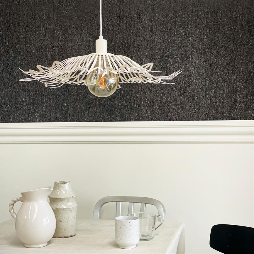 En hvid, dekorativ hængelampe svæver over et bord, hvor keramikkander, et glas og en keramikkop står; bagvæggen er mørk og tekstureret over en hvid, profileret liste.