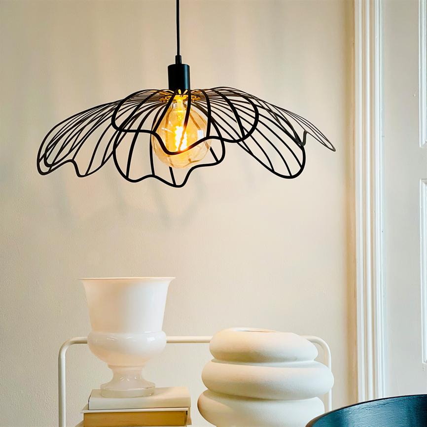 En sort, blomstret trådpendel hænger og udsender varmt glødelampe-lys over et bord med to hvide vaser foran en lys væg og vindueskarm.