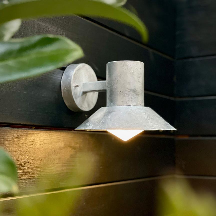 En metalvægslampe lyser nedad med et varmt skær, monteret på mørke træpaneler, omgivet af slørede grønne planteblade i forgrunden.