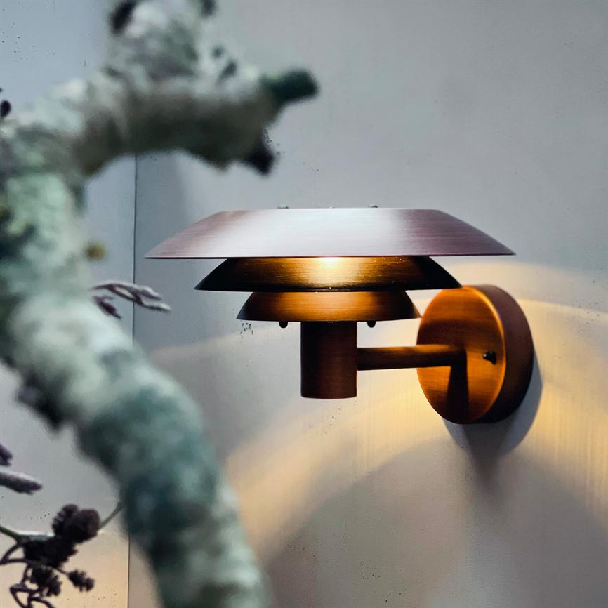 Væglampe i bronze; udsender varmt, blødt lys nedad og udad; monteret på en lys væg med en sløret, lavdækket gren i forgrunden.