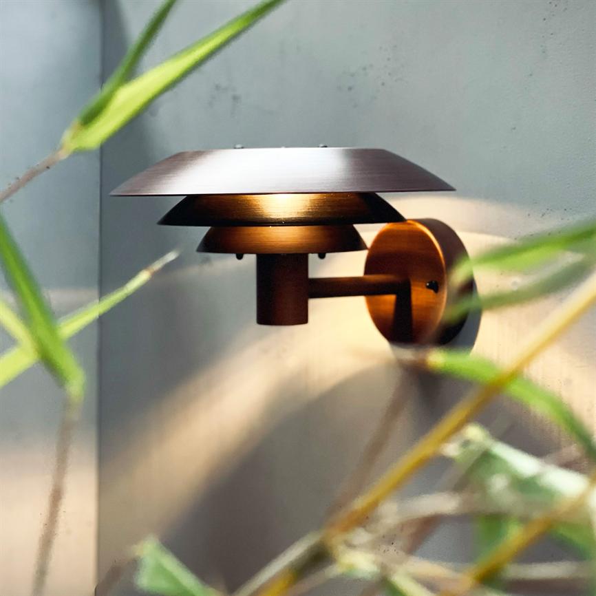 Væglampe udsender blødt varmt lys; bronzefarvet, lagdelt skærm monteret på rund vægbeslag; grønne planteblade i forgrunden, lyset kaster bløde skygger på grå væg.