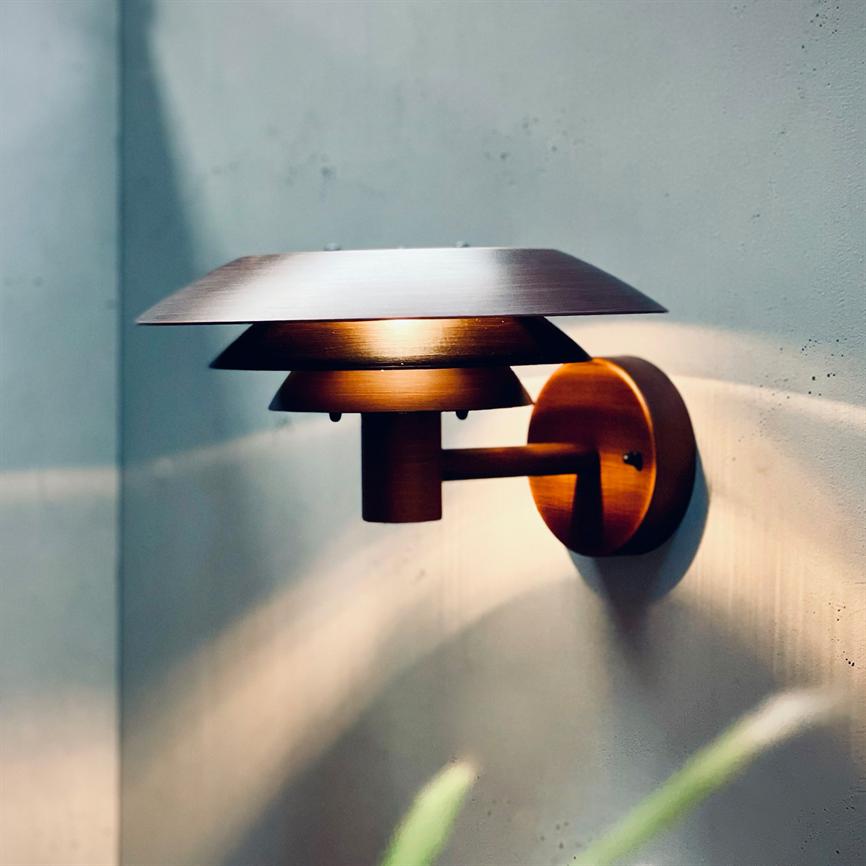 Væglampe i brunmetal kaster varmt lys gennem tre skærme, monteret på en betonvæg, hvilket skaber bløde lysstråler og skygger; en grøn plante er sløret i forgrunden.