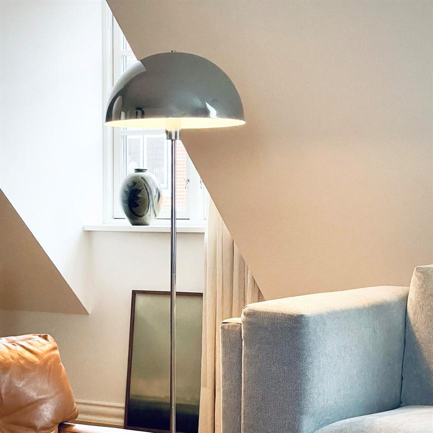 Gulvlampe lyser blødt; står foran et skråt vindue med en keramisk vase på karmen; omgivet af en lysegrå sofa og et brunt læderhjørne i et roligt, moderne stuehjørne.