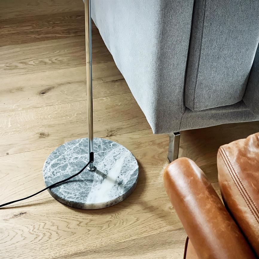 Gulvlampe med rund marmorfod står ved siden af en grå betrukket sofa; kromstang og sort ledning ligger på lyst trægulv, tæt på en brun læderstol.