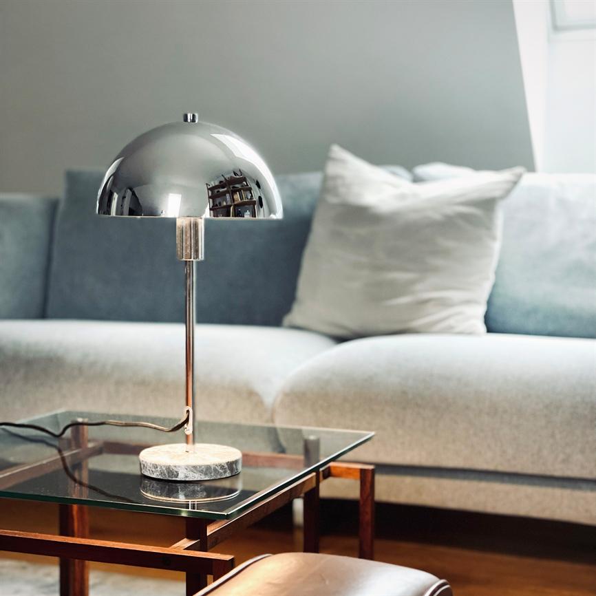 En halvkugleformet krom bordlampe står på et glast sidebord foran en lysegrå sofa med puder i en rolig, moderne stue.